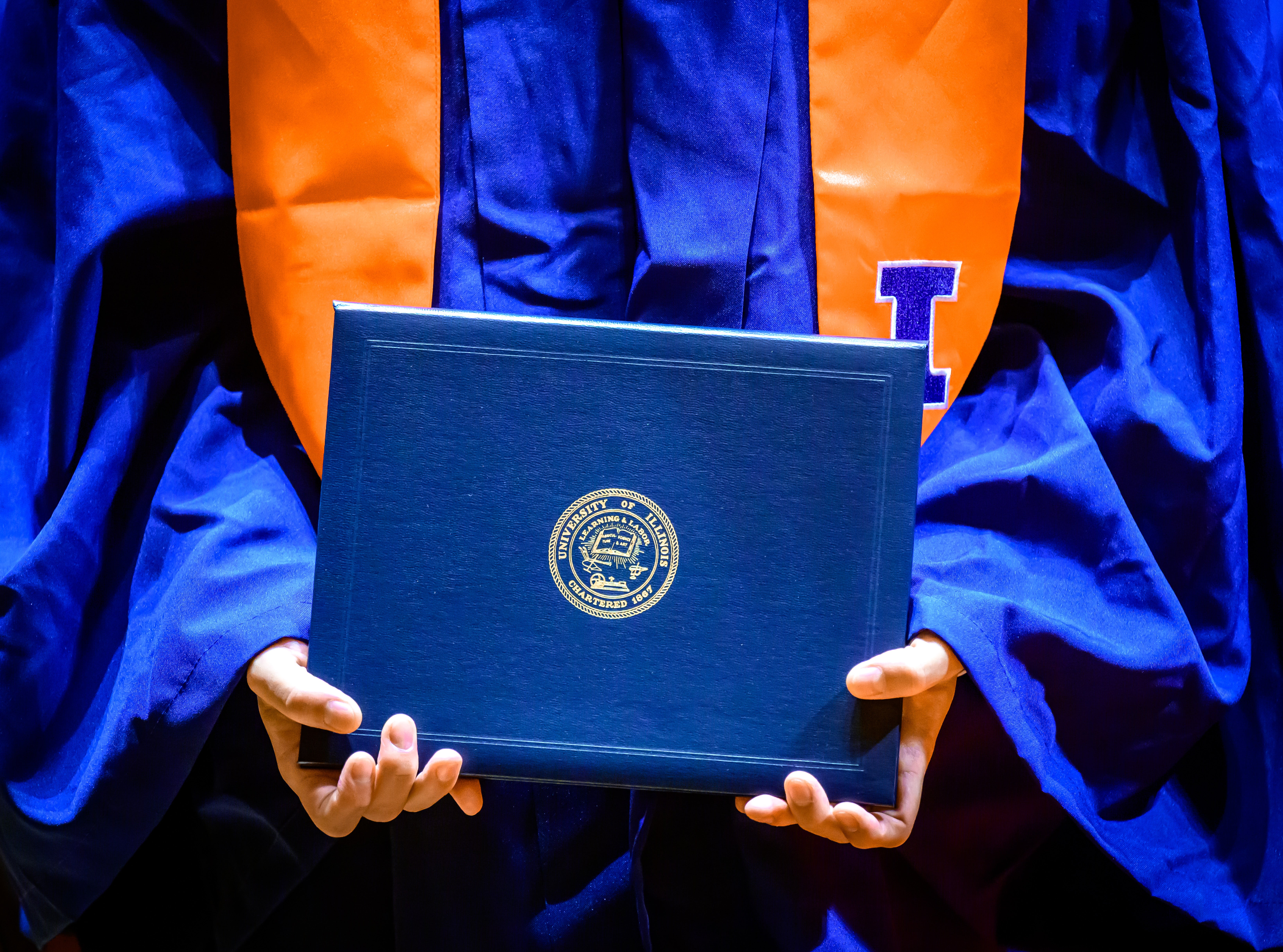 A student, in gown and stole, holds their diploma.