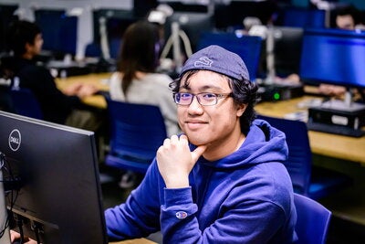 A student poses and smiles as he sits at a computer in a lab.