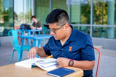A student in a blue Illini polo studies his textbook while sitting outside of the Siebel Center for Design.